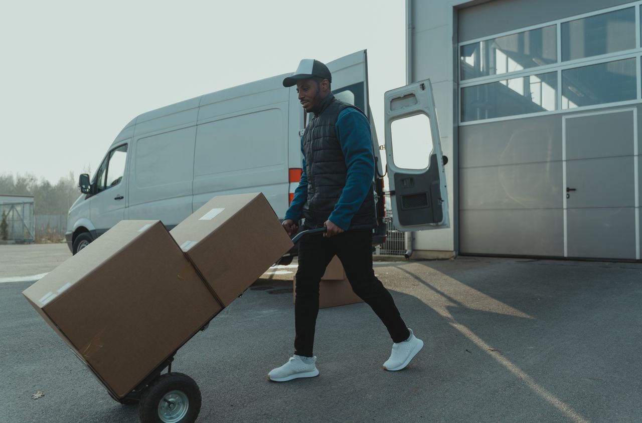 Courier pushing a trolley with boxes near a van at a warehouse, showcasing delivery services.